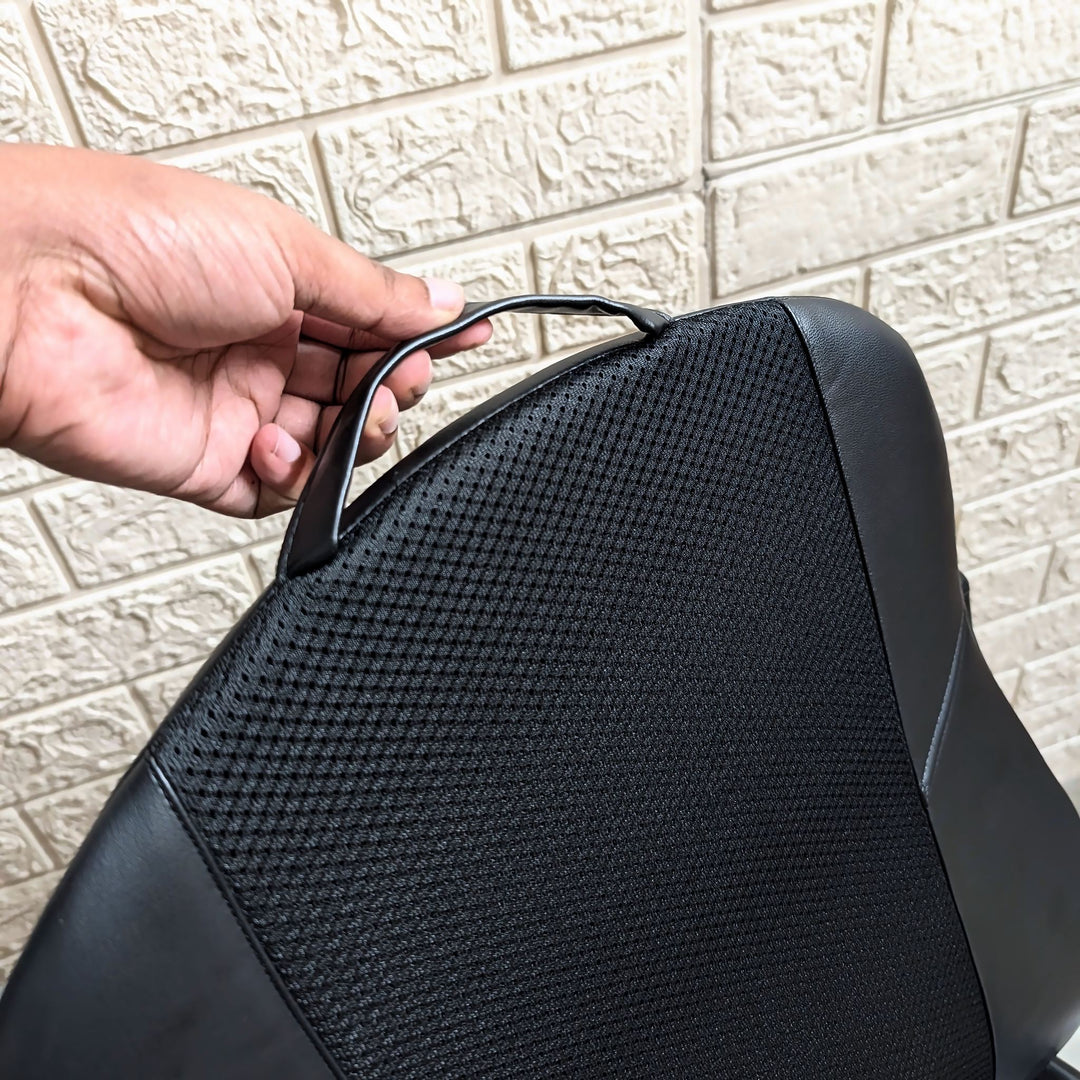 A close-up of a black mesh and faux-leather chair back cushion being held by a hand, with a light-colored textured brick wall in the background.