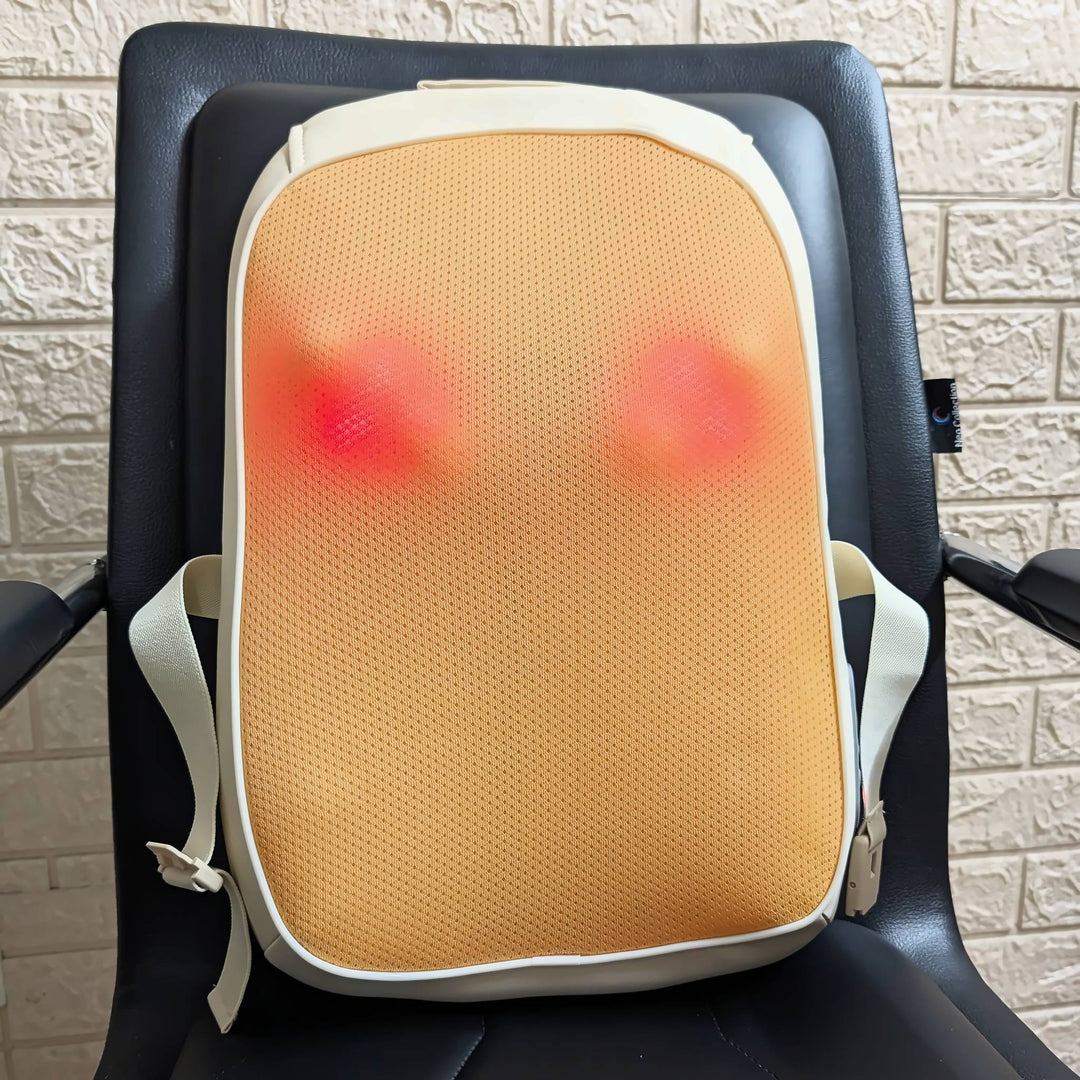 An electric hot massage lumbar pillow with a beige mesh body and white straps, resting on a black office chair in front of a light-colored brick wall.