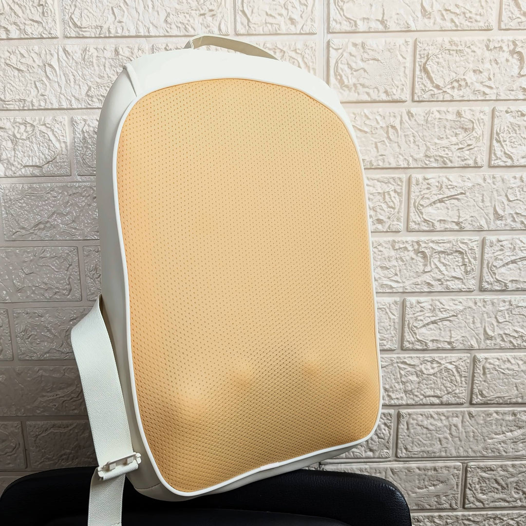 A white and beige ergonomic lumbar support pillow with a textured mesh backrest, resting on a black chair against a white brick wall.