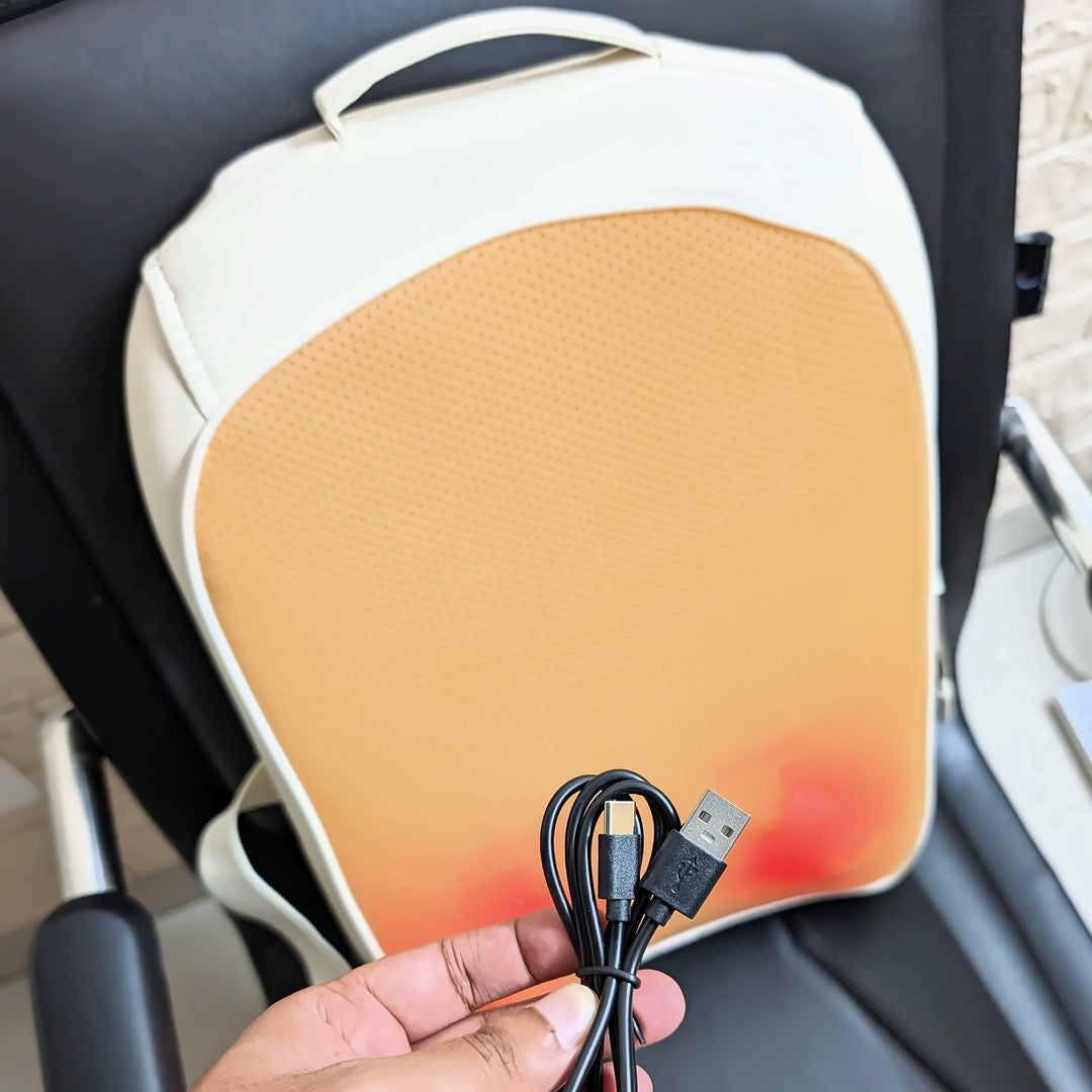 A close-up of a USB-controlled heated lumbar pillow, showing the black cable held by a hand in front of the white and orange cushion on a black office chair.